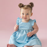 A light-skinned, blue-eyed baby with brown hair in two buns sits barefoot on a pale pink floor, wearing the Pearly Girly Flutter Sleeve Dress by Isobella and Chloe and smiling softly.