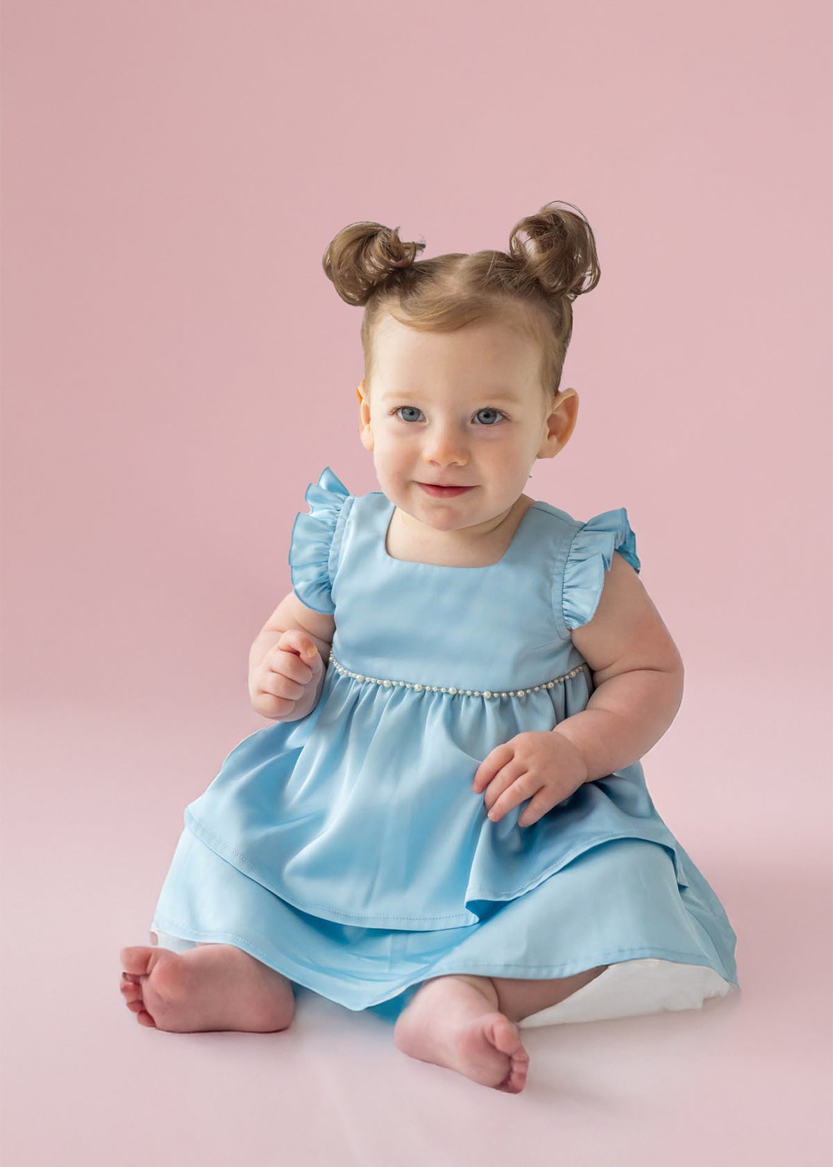 A light-skinned, blue-eyed baby with brown hair in two buns sits barefoot on a pale pink floor, wearing the Pearly Girly Flutter Sleeve Dress by Isobella and Chloe and smiling softly.