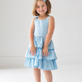A young girl with short light brown hair and a blue bow smiles in the Isobella and Chloe Pearly Girly Sleeveless Dress, featuring a ruffled skirt, and silver shoes, against a plain white background.