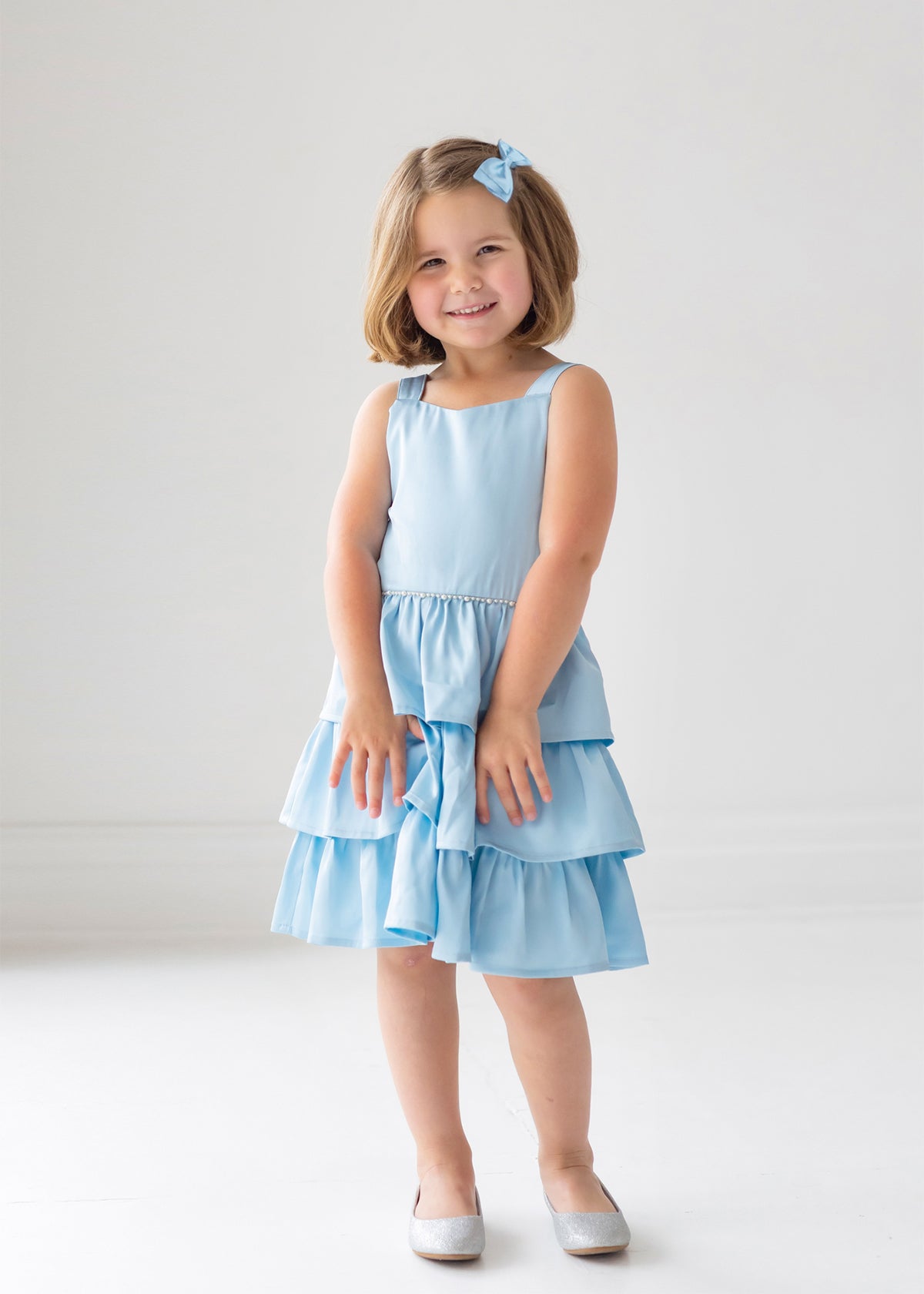 A young girl with short light brown hair and a blue bow smiles in the Isobella and Chloe Pearly Girly Sleeveless Dress, featuring a ruffled skirt, and silver shoes, against a plain white background.