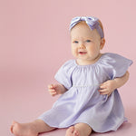 A baby sits barefoot on a pink background, smiling softly in the Isobella and Chloe Diamond Dazzle Puff Sleeve Purple Dress with a matching bow headband.