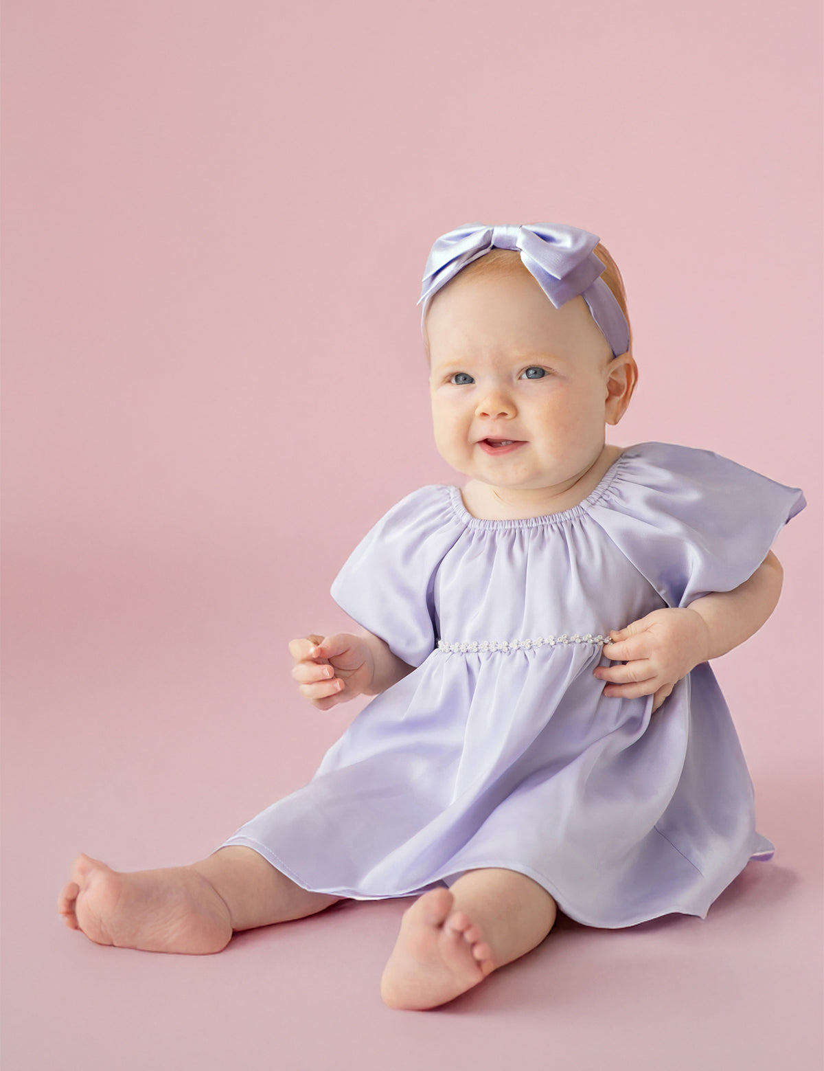 A baby sits barefoot on a pink background, smiling softly in the Isobella and Chloe Diamond Dazzle Puff Sleeve Purple Dress with a matching bow headband.