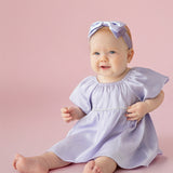 A baby sits barefoot on a pink background, smiling softly in the Isobella and Chloe Diamond Dazzle Puff Sleeve Purple Dress with a matching bow headband.