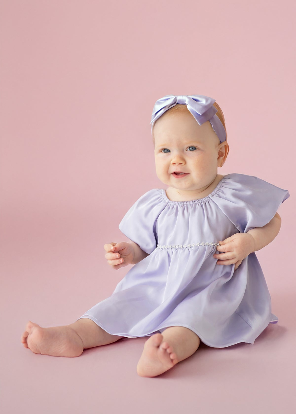 A baby sits barefoot on a pink background, smiling softly in the Isobella and Chloe Diamond Dazzle Puff Sleeve Purple Dress with a matching bow headband.