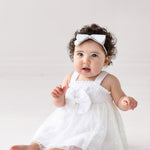 A baby with curly hair wears the Isobella and Chloe Birds of a Feather Sleeveless Dress and a white headband, sitting barefoot on a light floor and looking up with an open mouth—perfect for milestone photos.