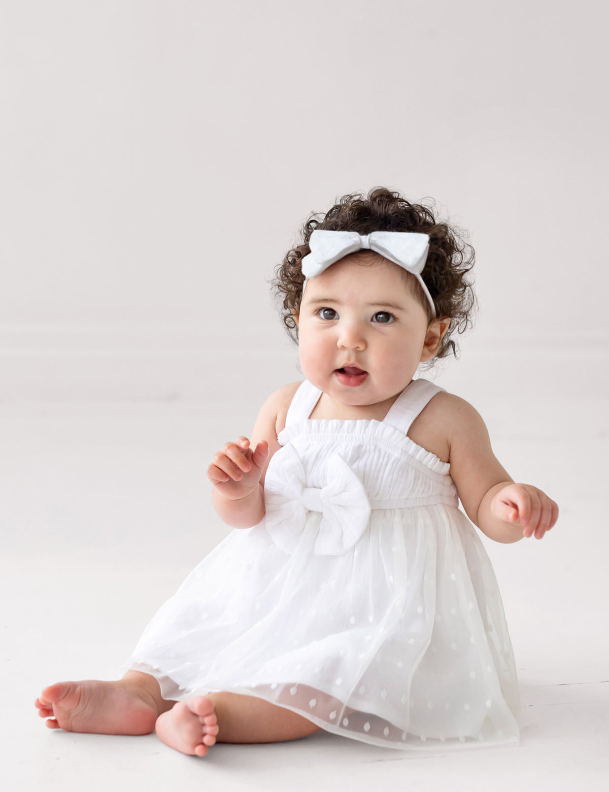 A baby with curly hair wears the Isobella and Chloe Birds of a Feather Sleeveless Dress and a white headband, sitting barefoot on a light floor and looking up with an open mouth—perfect for milestone photos.