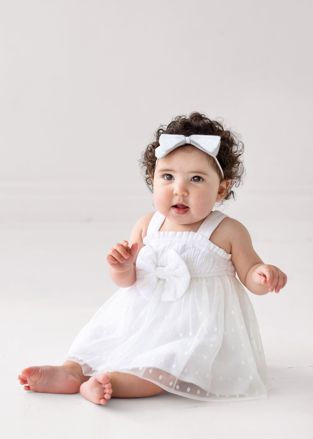 A baby with curly hair wears the Isobella and Chloe Birds of a Feather Sleeveless Dress and a white headband, sitting barefoot on a light floor and looking up with an open mouth—perfect for milestone photos.