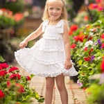 A young girl wearing the Isobella and Chloe Birds of a Feather Cap Sleeve Dress and sandals smiles while standing on a brick path surrounded by vibrant red and pink garden flowers.