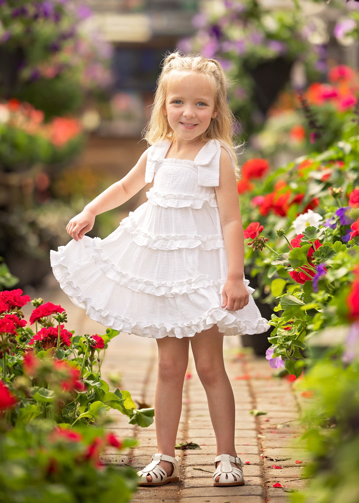 A young girl wearing the Isobella and Chloe Birds of a Feather Cap Sleeve Dress and sandals smiles while standing on a brick path surrounded by vibrant red and pink garden flowers.