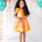 A young girl with long curly hair, wearing an Isobella and Chloe Papaya Paradise Sleeveless Dress and gold shoes, smiles among teal and pink balloons with orange confetti against a white background.