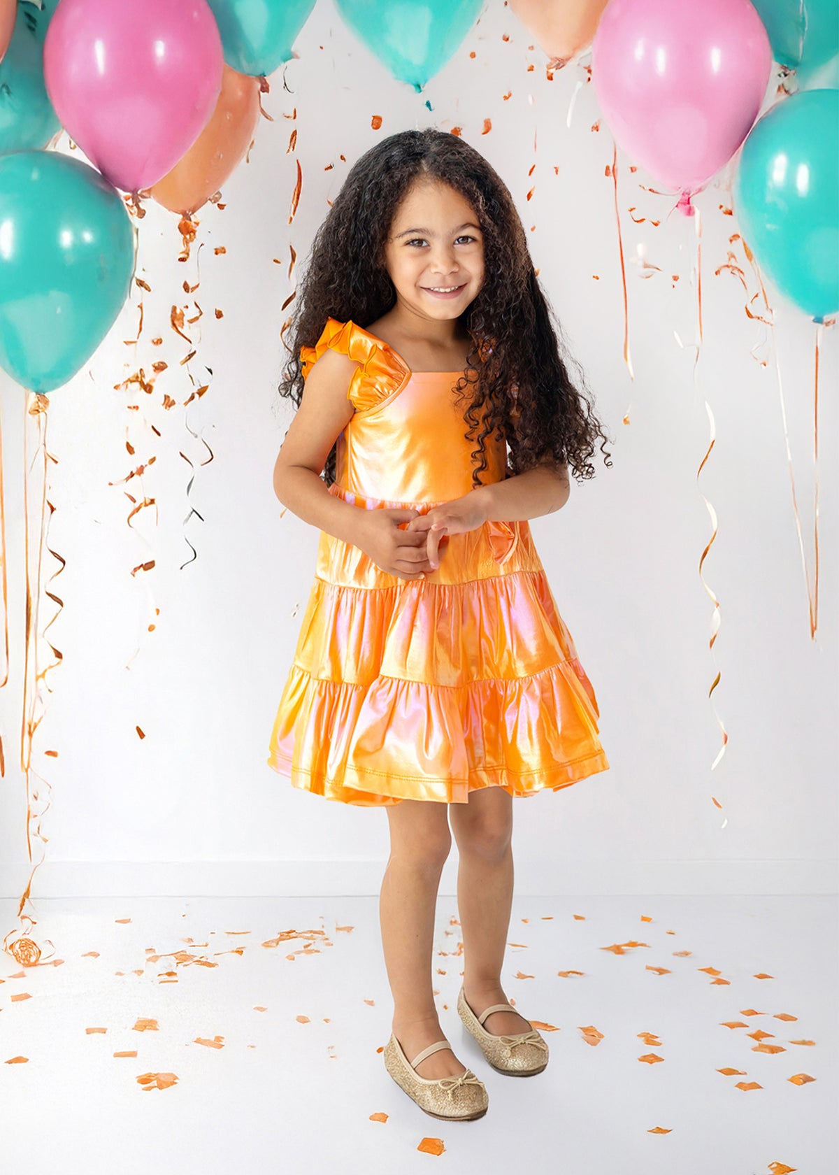 A young girl with long curly hair, wearing an Isobella and Chloe Papaya Paradise Sleeveless Dress and gold shoes, smiles among teal and pink balloons with orange confetti against a white background.