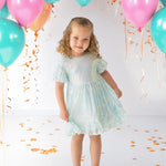 A young girl beams in an Isobella and Chloe Ocean Shimmer Short Sleeve Dress, surrounded by teal, pink, and orange balloons and confetti—her curly hair and silver shoes complete a festive look perfect for any special occasion.