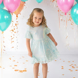 A young girl beams in an Isobella and Chloe Ocean Shimmer Short Sleeve Dress, surrounded by teal, pink, and orange balloons and confetti—her curly hair and silver shoes complete a festive look perfect for any special occasion.