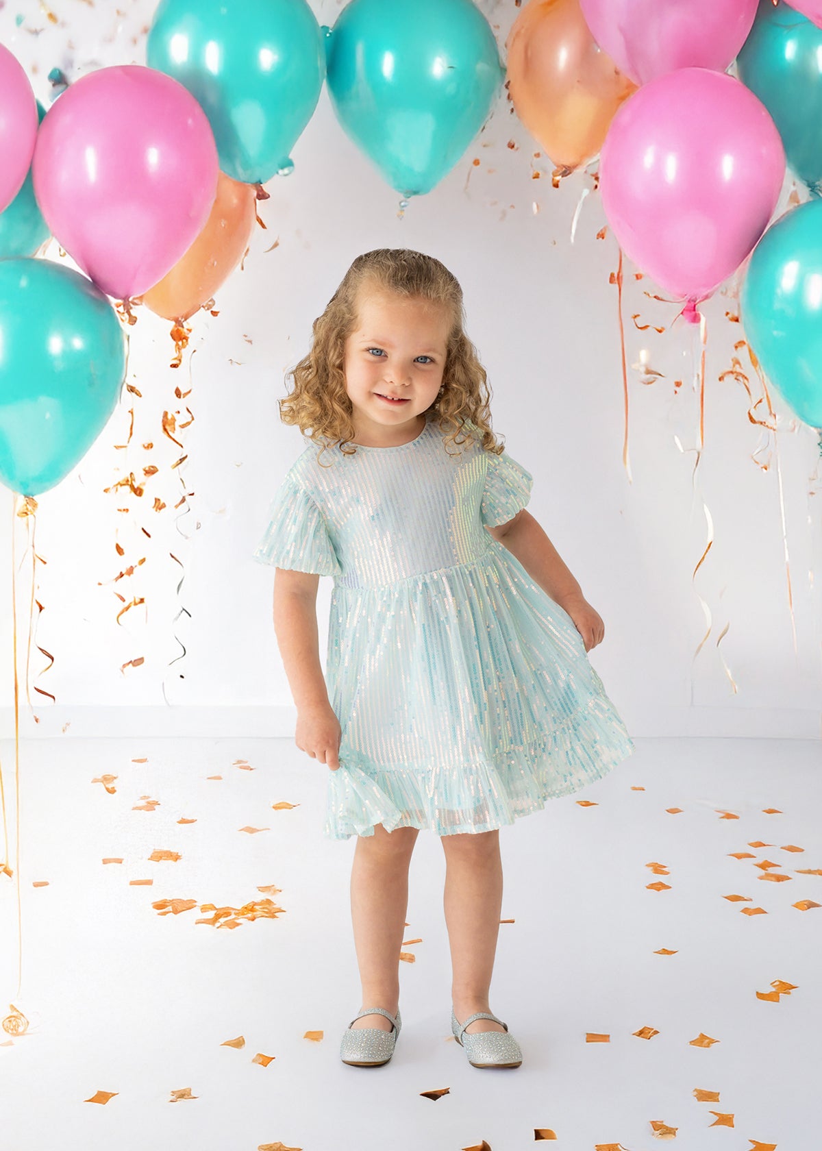 A young girl beams in an Isobella and Chloe Ocean Shimmer Short Sleeve Dress, surrounded by teal, pink, and orange balloons and confetti—her curly hair and silver shoes complete a festive look perfect for any special occasion.