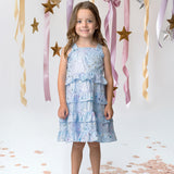A young girl in an Isobella and Chloe Starry Glitters Tank Dress smiles on a white floor with rose gold confetti, surrounded by gold stars and pastel ribbons—perfect as a magical birthday or girls party dress.