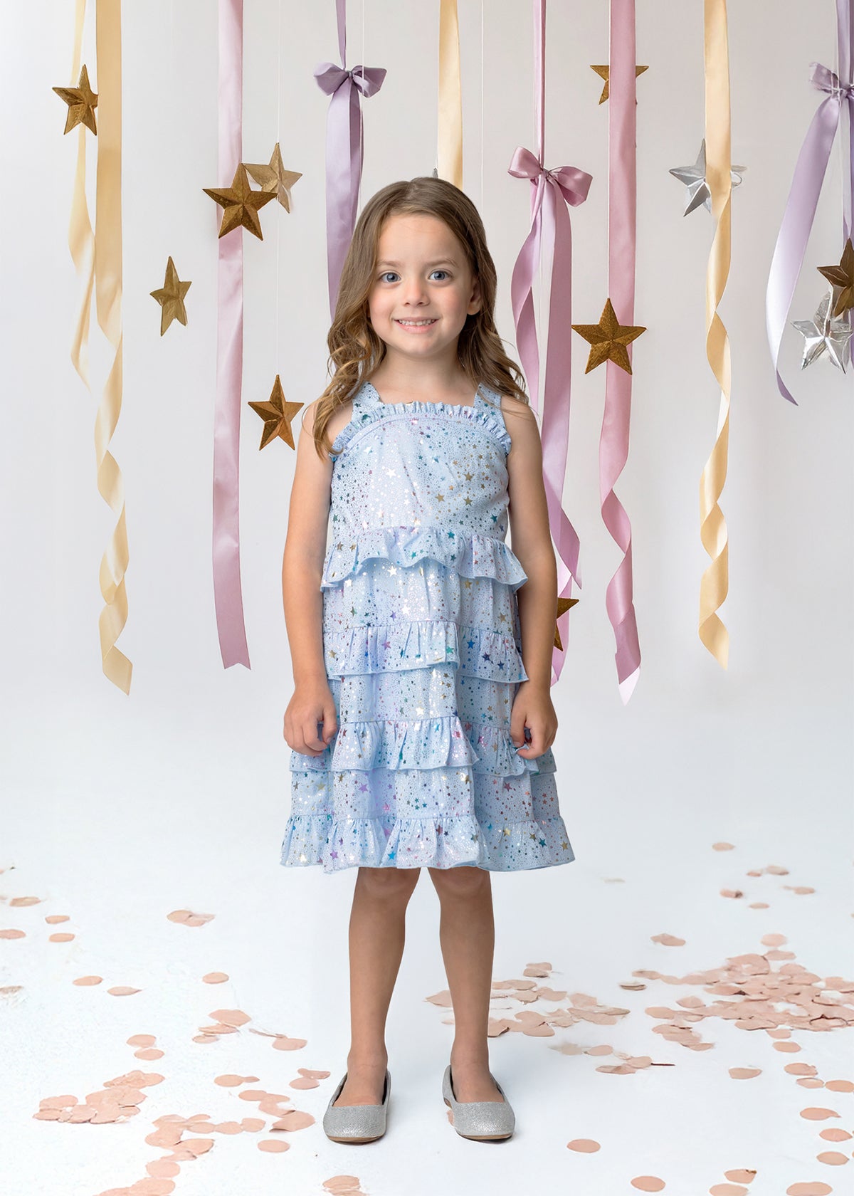 A young girl in an Isobella and Chloe Starry Glitters Tank Dress smiles on a white floor with rose gold confetti, surrounded by gold stars and pastel ribbons—perfect as a magical birthday or girls party dress.