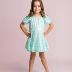 Smiling young girl with long brown hair poses hands on hips in an Isobella and Chloe Teal Twinkle Short Sleeve Dress and beige shoes, set against a light pink background.