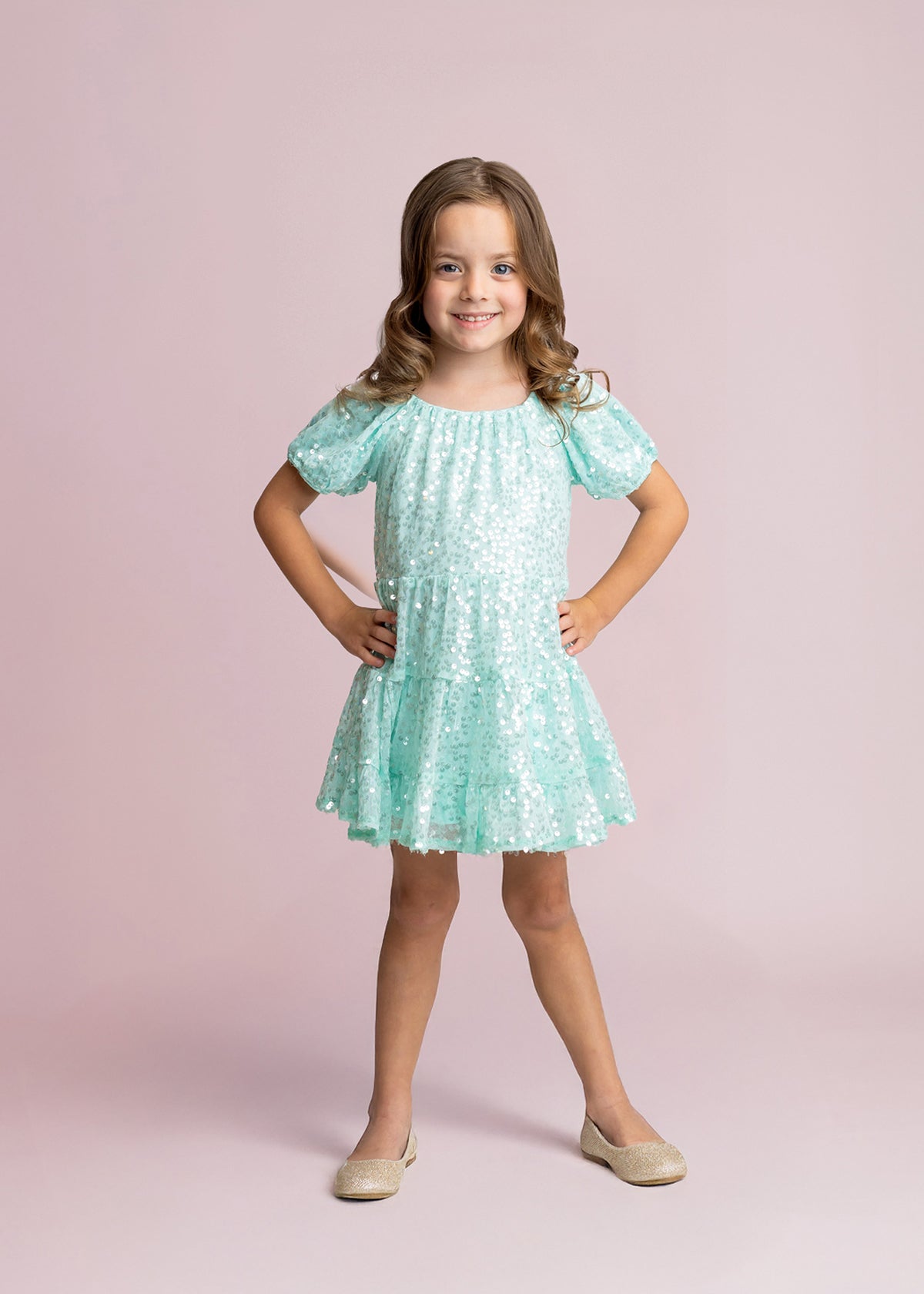 Smiling young girl with long brown hair poses hands on hips in an Isobella and Chloe Teal Twinkle Short Sleeve Dress and beige shoes, set against a light pink background.