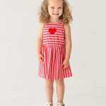 A young girl with curly hair smiles on a white platform, wearing the Mabel and Honey Sugar Sweet Sleeveless Dress with red and pink stripes, a red heart on the chest, and white sandals against a light background.