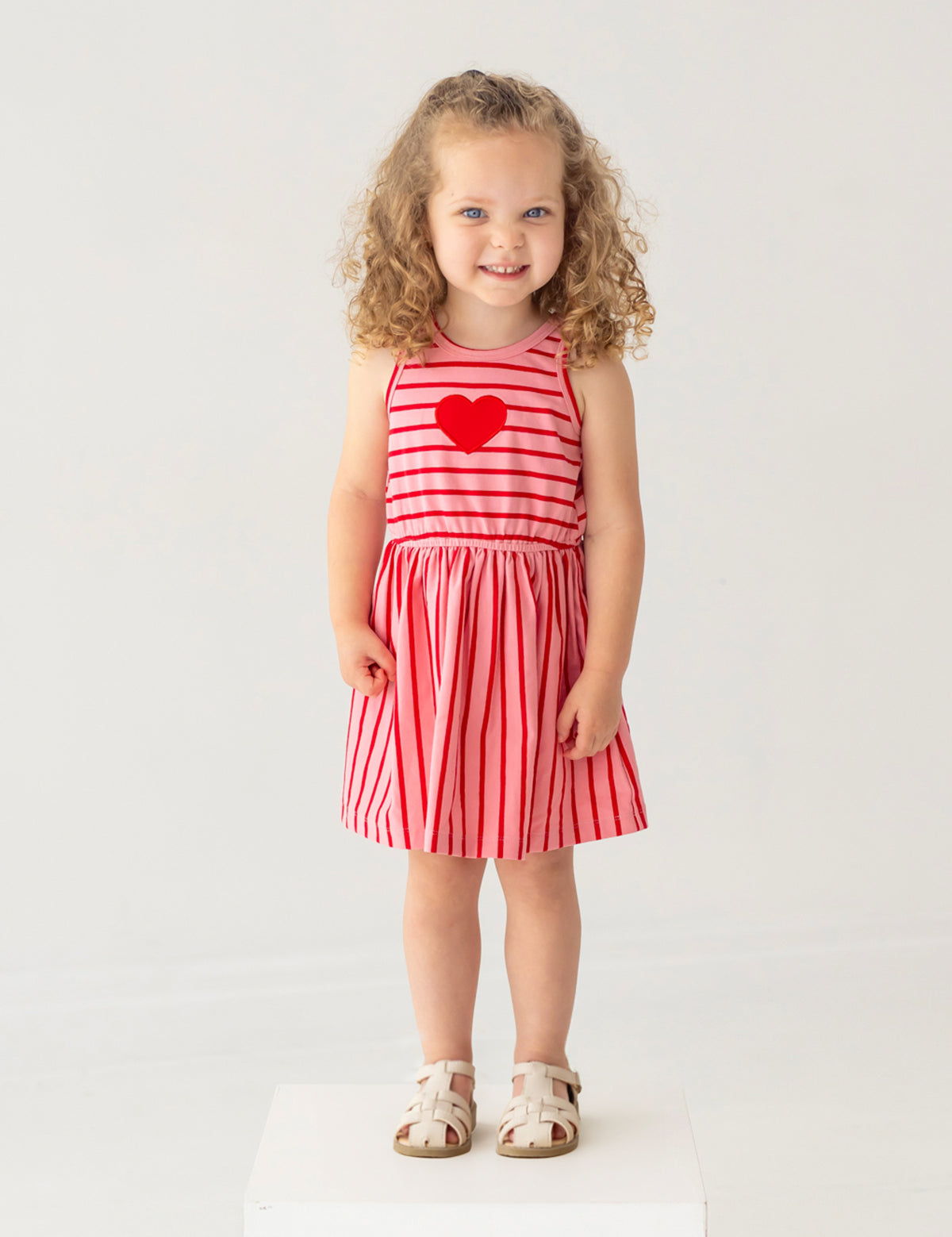 A young girl with curly hair smiles on a white platform, wearing the Mabel and Honey Sugar Sweet Sleeveless Dress with red and pink stripes, a red heart on the chest, and white sandals against a light background.