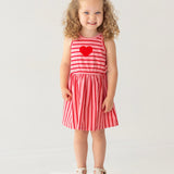 A young girl with curly hair smiles on a white platform, wearing the Mabel and Honey Sugar Sweet Sleeveless Dress with red and pink stripes, a red heart on the chest, and white sandals against a light background.
