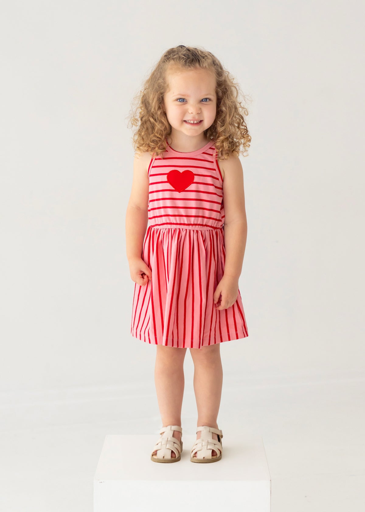 A young girl with curly hair smiles on a white platform, wearing the Mabel and Honey Sugar Sweet Sleeveless Dress with red and pink stripes, a red heart on the chest, and white sandals against a light background.