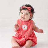 A curly-haired baby wears a Mabel and Honey Striped Blossom Sleeveless Romper with a matching headband, sitting barefoot on a white floor and gazing slightly upward.