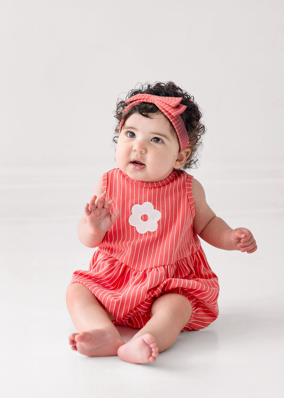 A curly-haired baby wears a Mabel and Honey Striped Blossom Sleeveless Romper with a matching headband, sitting barefoot on a white floor and gazing slightly upward.