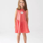 A young girl with shoulder-length brown hair smiles in a Mabel and Honey Striped Blossom Sleeveless Dress, featuring red and white stripes with a flower applique, paired with white sandals against a plain white background.