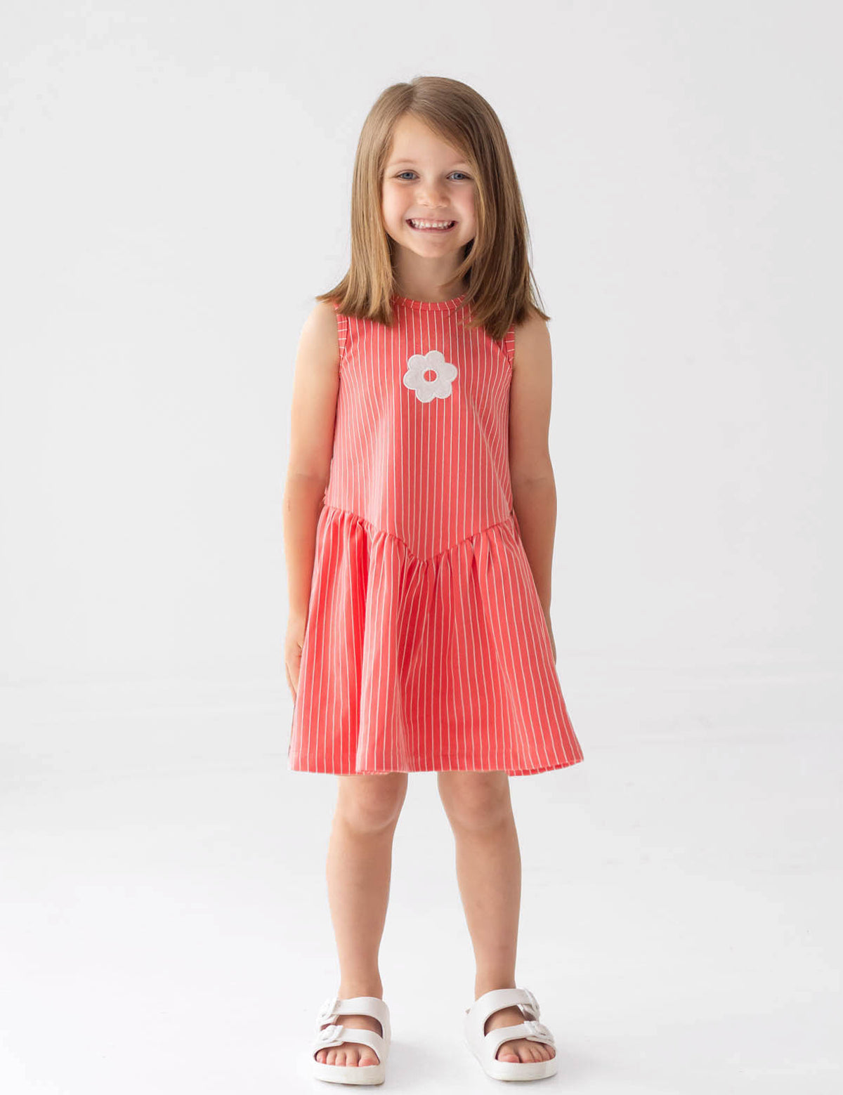 A young girl with shoulder-length brown hair smiles in a Mabel and Honey Striped Blossom Sleeveless Dress, featuring red and white stripes with a flower applique, paired with white sandals against a plain white background.