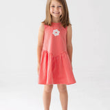 A young girl with shoulder-length brown hair smiles in a Mabel and Honey Striped Blossom Sleeveless Dress, featuring red and white stripes with a flower applique, paired with white sandals against a plain white background.