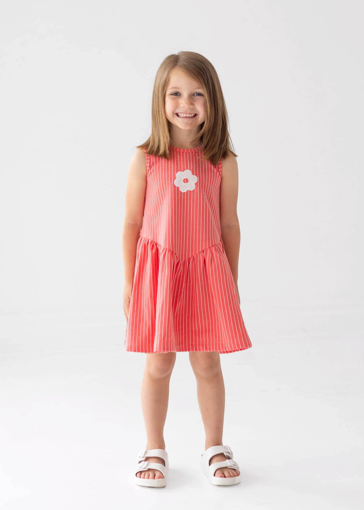 A young girl with shoulder-length brown hair smiles in a Mabel and Honey Striped Blossom Sleeveless Dress, featuring red and white stripes with a flower applique, paired with white sandals against a plain white background.