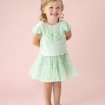 A young blonde girl smiles against a pale pink backdrop, wearing the Isobella and Chloe Alice Short Ruffle Sleeve Top in mint green, styled as a dress with front bows, a ruffled skirt, and clear sandals. Her hands rest behind her back.