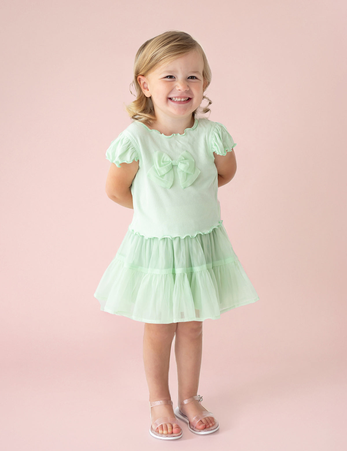 A young blonde girl smiles against a pale pink backdrop, wearing the Isobella and Chloe Alice Short Ruffle Sleeve Top in mint green, styled as a dress with front bows, a ruffled skirt, and clear sandals. Her hands rest behind her back.