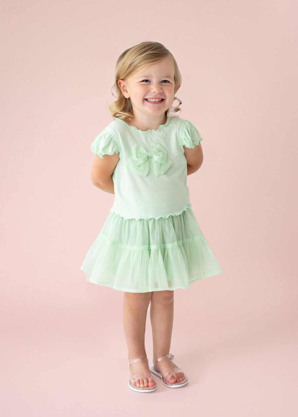A young blonde girl smiles against a pale pink backdrop, wearing the Isobella and Chloe Alice Short Ruffle Sleeve Top in mint green, styled as a dress with front bows, a ruffled skirt, and clear sandals. Her hands rest behind her back.