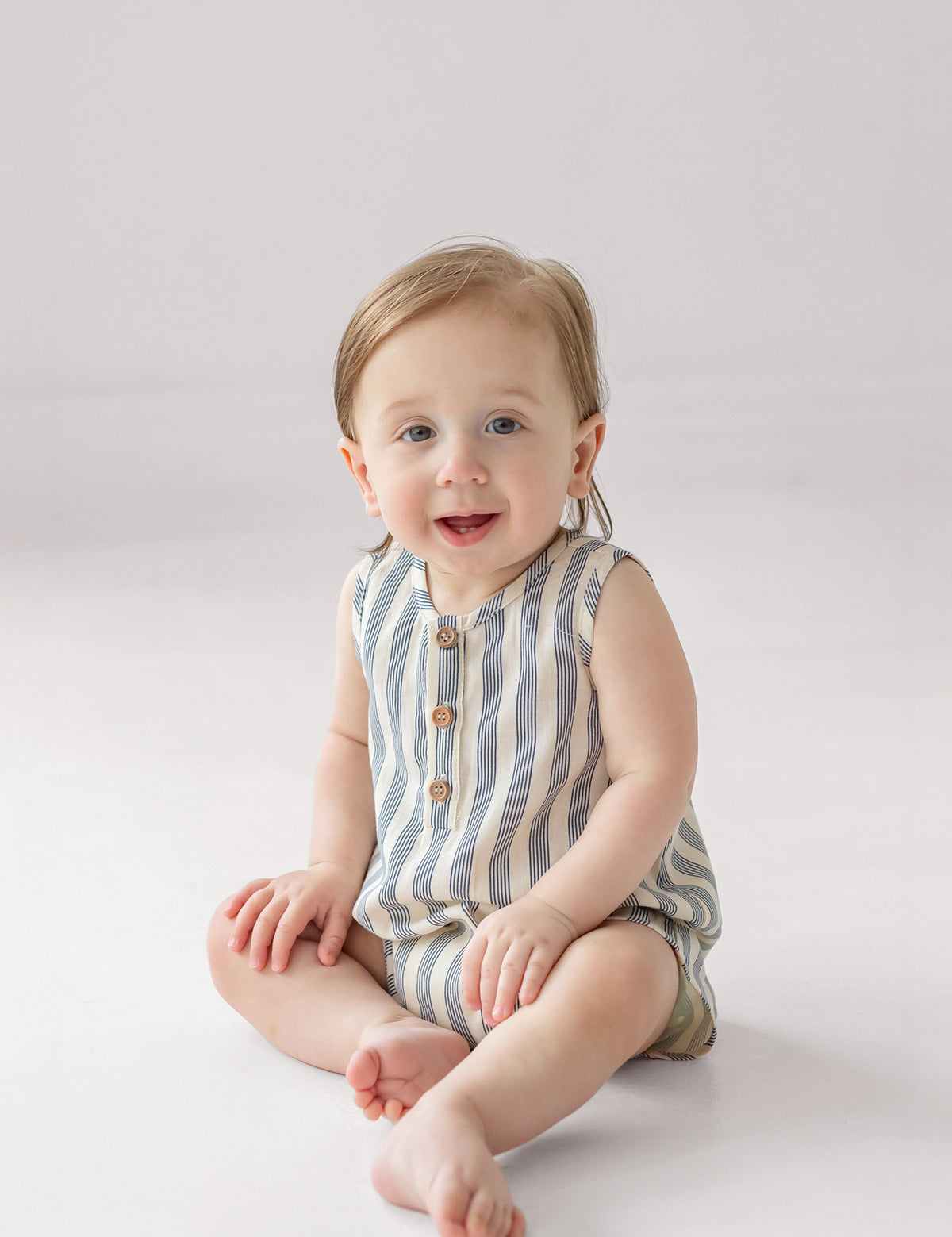 A smiling baby with light hair sits on the floor wearing the Beckett and Bear Knox Sleeveless Romper, which features stripes and front buttons, against a plain light background.
