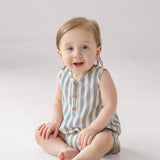 A smiling baby with light hair sits on the floor wearing the Beckett and Bear Knox Sleeveless Romper, which features stripes and front buttons, against a plain light background.