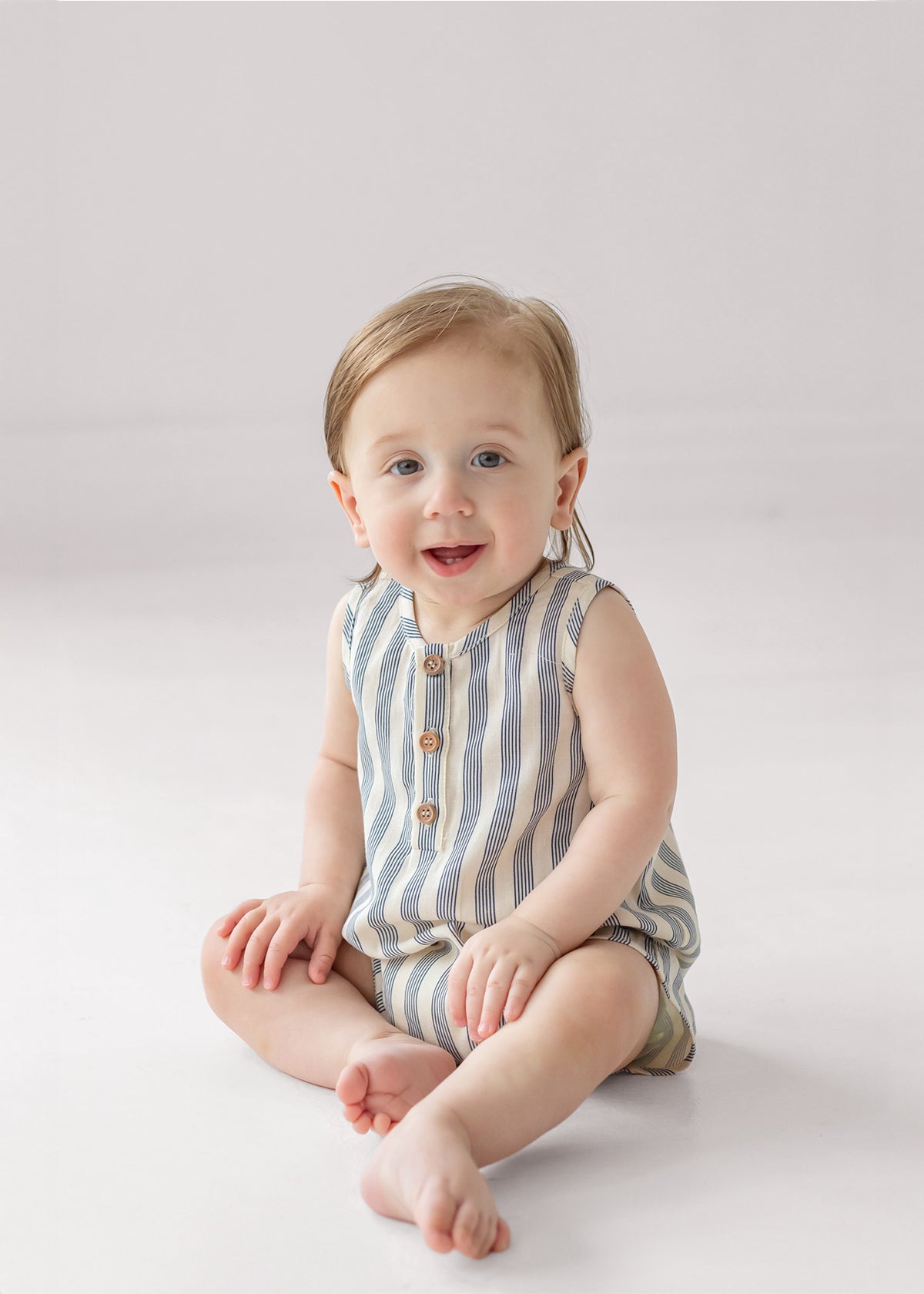 A smiling baby with light hair sits on the floor wearing the Beckett and Bear Knox Sleeveless Romper, which features stripes and front buttons, against a plain light background.