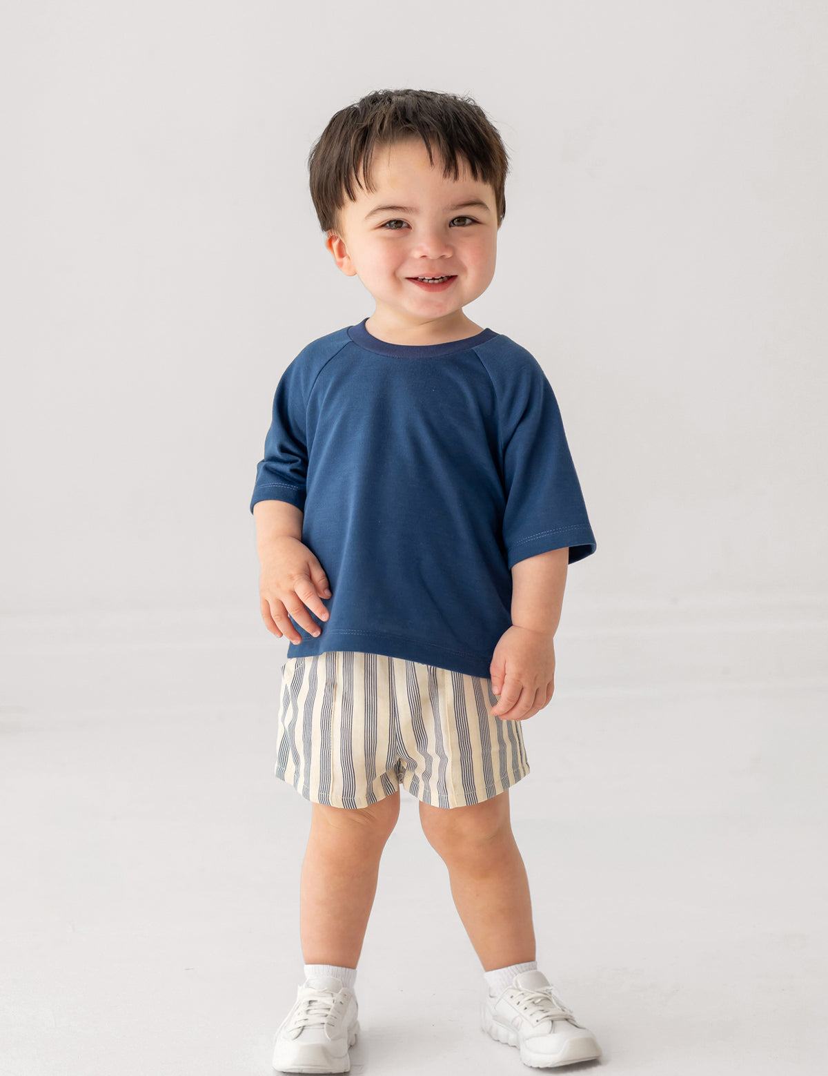 A smiling toddler wears Beckett and Bear's Knox Shorts Short, a blue-striped pair, with a blue short-sleeved shirt and white sneakers, standing against a plain light background.