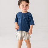 A smiling toddler wears Beckett and Bear's Knox Shorts Short, a blue-striped pair, with a blue short-sleeved shirt and white sneakers, standing against a plain light background.