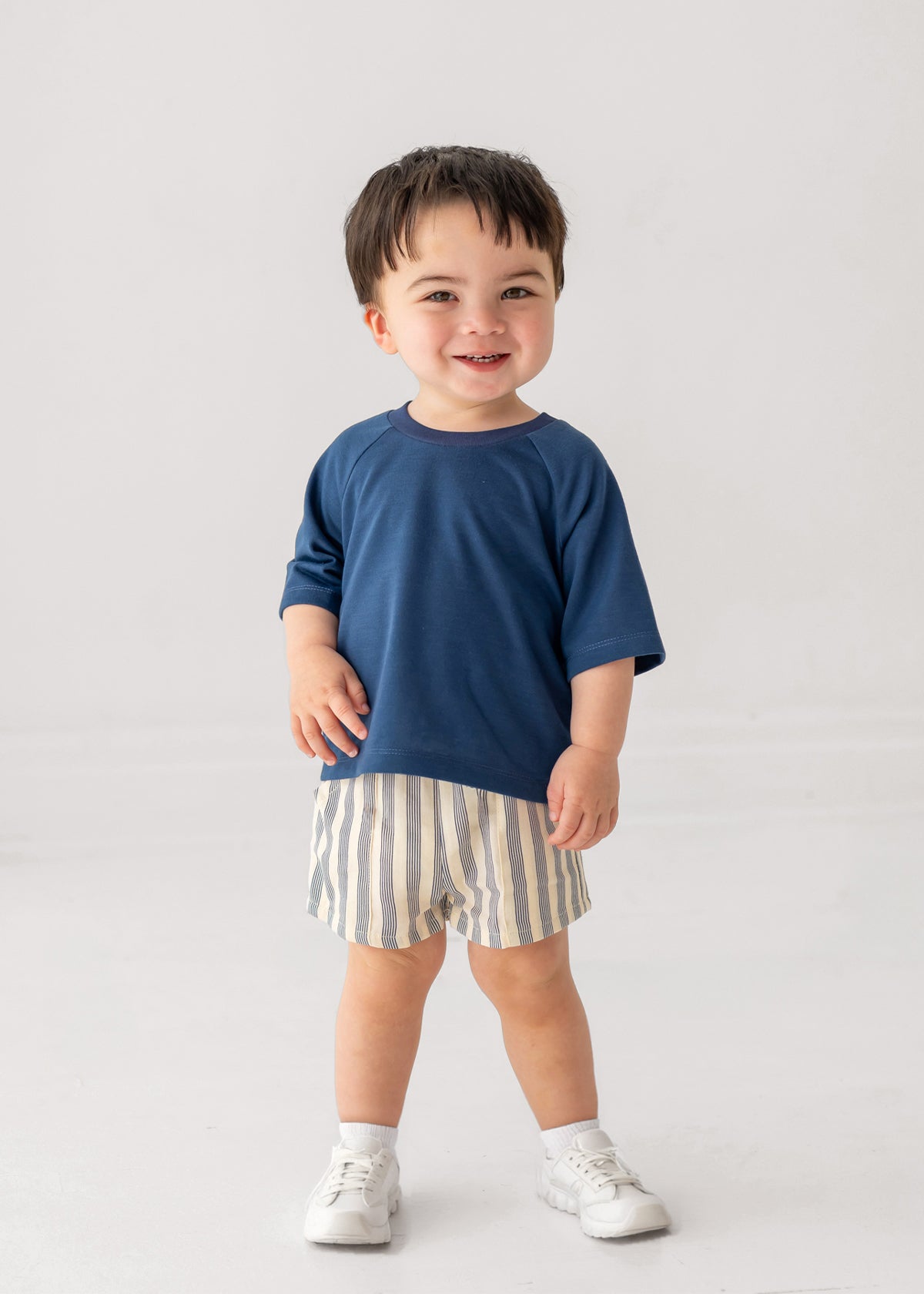 A smiling toddler wears Beckett and Bear's Knox Shorts Short, a blue-striped pair, with a blue short-sleeved shirt and white sneakers, standing against a plain light background.