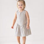 A smiling toddler with light brown hair stands on a wooden box, wearing a Beckett and Bear Knox Sleeveless Dress in blue and white stripes with beige sandals against a plain light background.