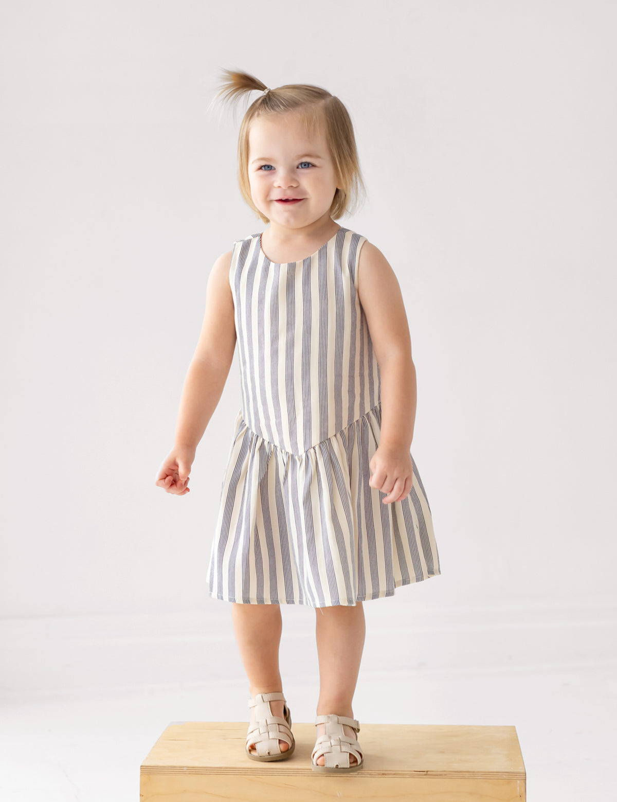 A smiling toddler with light brown hair stands on a wooden box, wearing a Beckett and Bear Knox Sleeveless Dress in blue and white stripes with beige sandals against a plain light background.