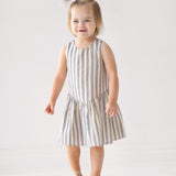 A smiling toddler with light brown hair stands on a wooden box, wearing a Beckett and Bear Knox Sleeveless Dress in blue and white stripes with beige sandals against a plain light background.