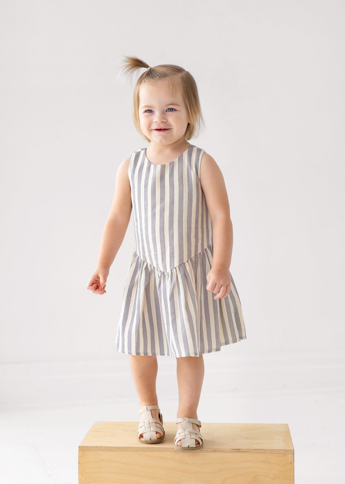 A smiling toddler with light brown hair stands on a wooden box, wearing a Beckett and Bear Knox Sleeveless Dress in blue and white stripes with beige sandals against a plain light background.