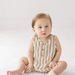 A baby with light brown hair and blue eyes sits on a white surface, wearing a Beckett and Bear Nash Sleeveless Romper made from breathable fabric, looking up with a calm expression.