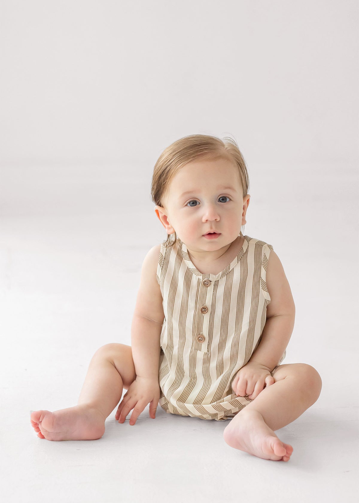 A baby with light brown hair and blue eyes sits on a white surface, wearing a Beckett and Bear Nash Sleeveless Romper made from breathable fabric, looking up with a calm expression.