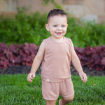 A smiling toddler stands barefoot on green grass, wearing the Beckett and Bear Dustin Sleeveless Two Piece Set. Bushes and a stone wall create a charming backdrop for this adorable kids’ outfit.