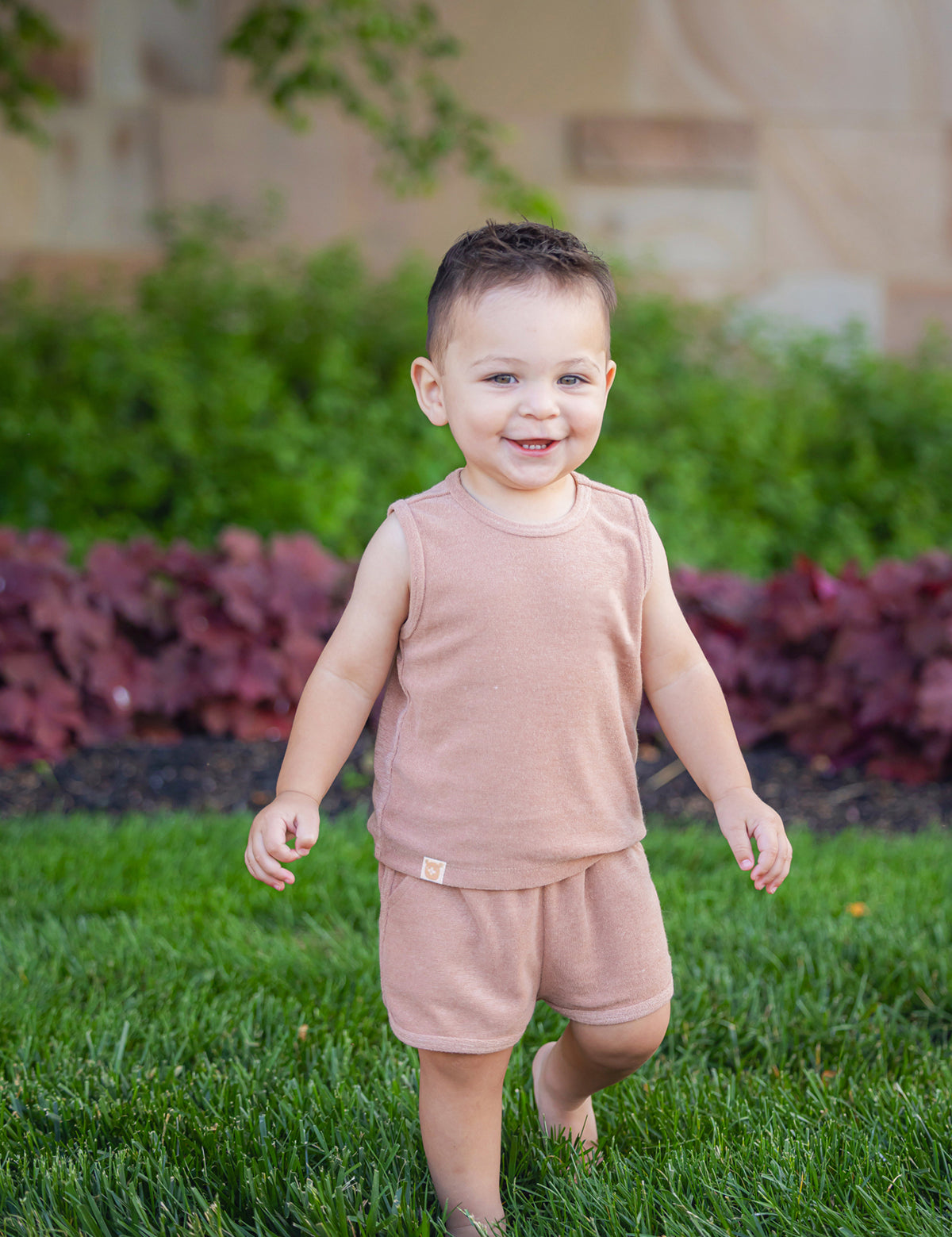 A smiling toddler stands barefoot on green grass, wearing the Beckett and Bear Dustin Sleeveless Two Piece Set. Bushes and a stone wall create a charming backdrop for this adorable kids’ outfit.