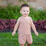 A smiling toddler stands barefoot on green grass, wearing the Beckett and Bear Dustin Sleeveless Two Piece Set. Bushes and a stone wall create a charming backdrop for this adorable kids’ outfit.